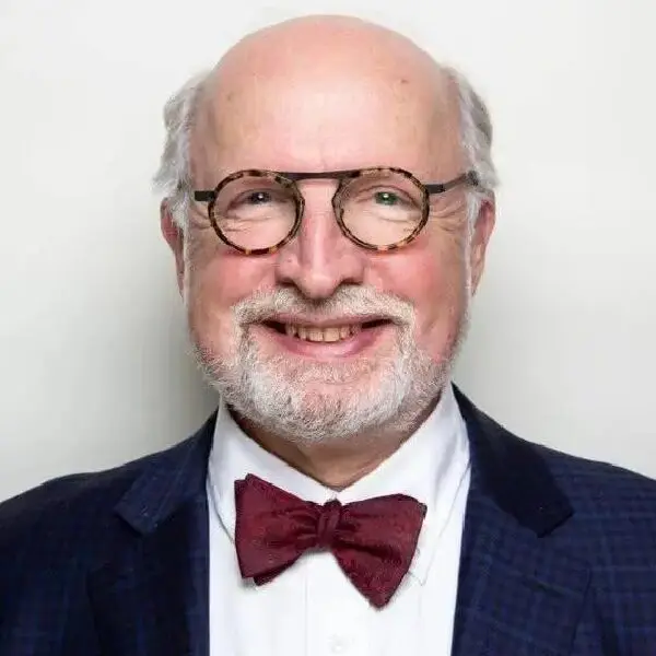 A man with glasses and bow tie in front of a white wall.